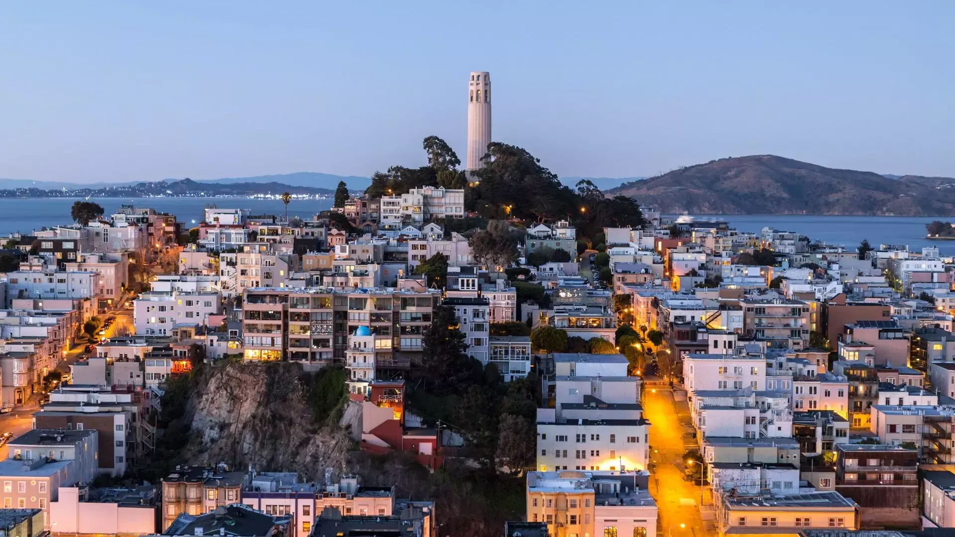 San Franciscos Coit Tower in der Abenddämmerung, mit beleuchteten Straßen davor und der Bucht von San Francisco dahinter.